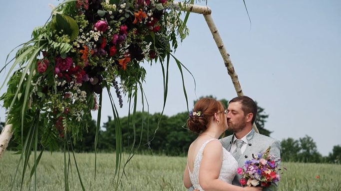 Brautpaar küsst sich unter einem blühenden Hochzeitsbogen in einer Wiese.