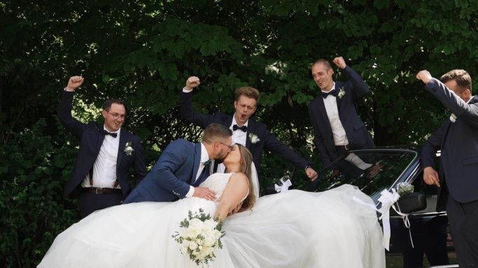 Braut und Bräutigam küssen sich, umgeben von freudigen Gästen in festlicher Kleidung.